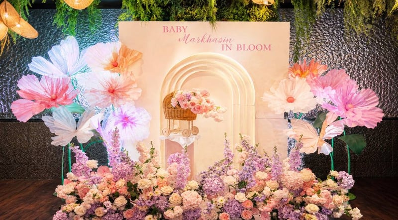 Cream arch backdrop with a “Baby in Bloom” sign, giant pastel paper flowers on both sides, a wicker pram and lavender and rose florals