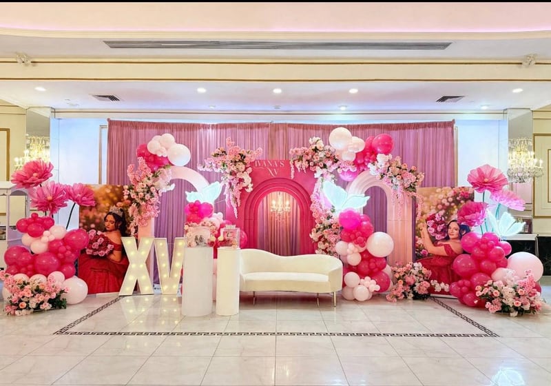 Wide view of a ballroom styled in hot pink, with a central arch backdrop, matching side arches, balloon columns, marquee letters and a cream sofa