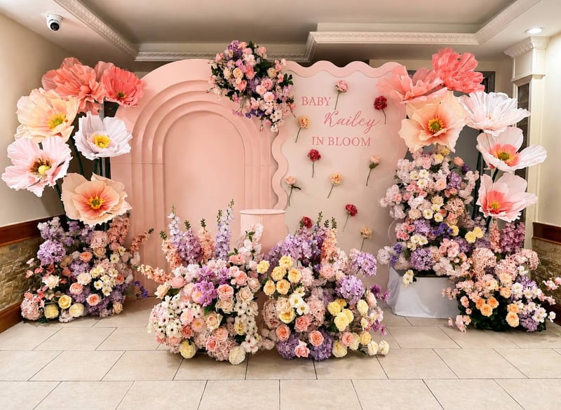 Peach arched backdrop with an “In Bloom” sign, oversized paper flowers and mounds of pink and lavender florals