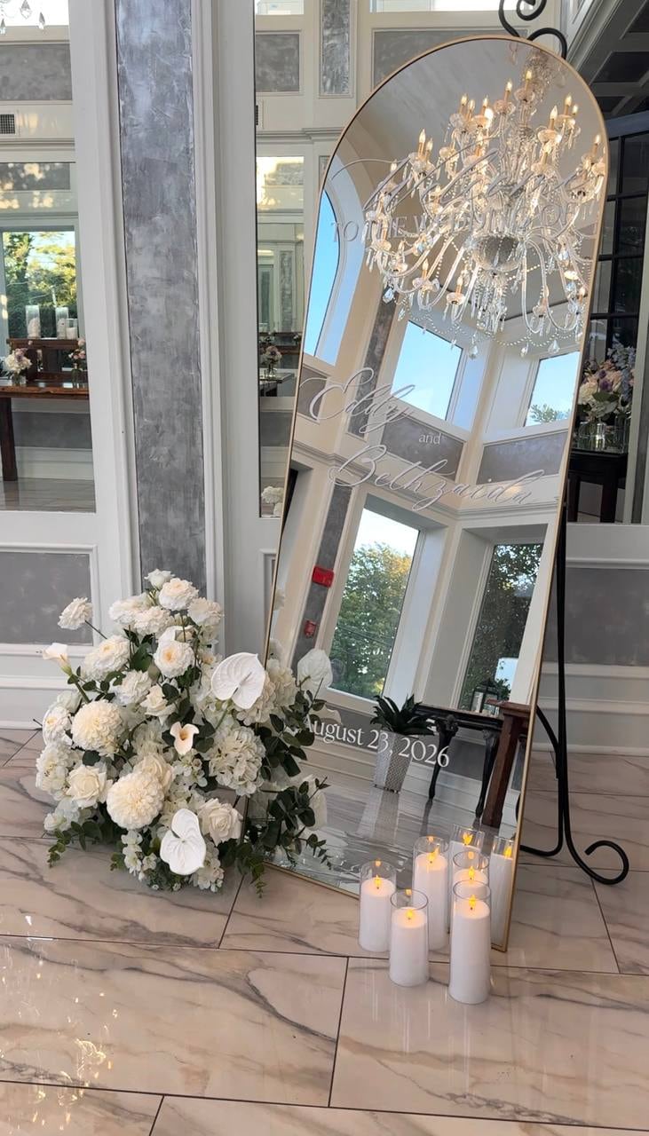 Tall arched mirror reflecting a chandelier, with a white floral arrangement and pillar candles at its base