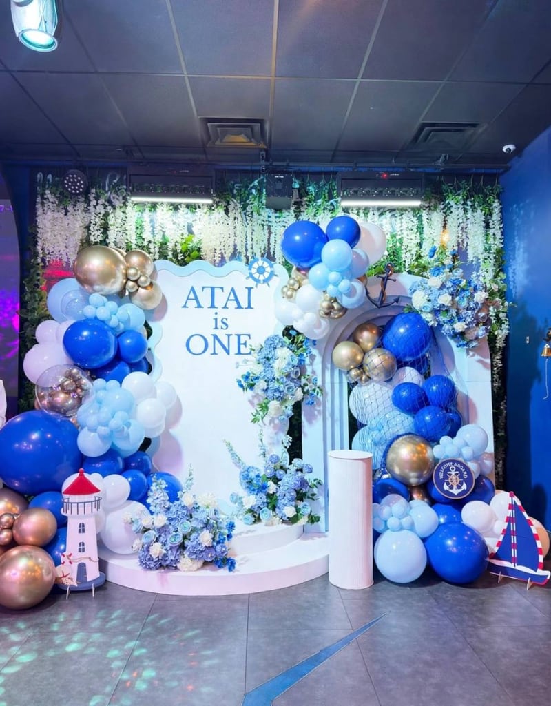 Blue, navy and gold balloons with white florals around a nautical first-birthday backdrop with a lighthouse prop