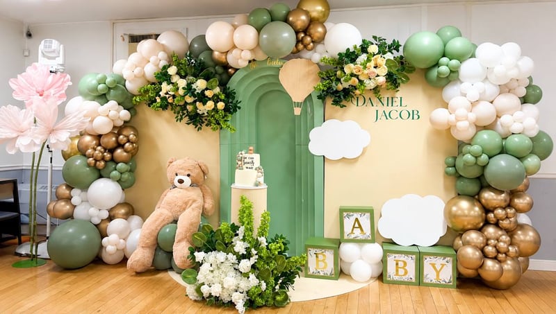 Sage green, cream and gold balloon arch with greenery, alphabet blocks and a large teddy bear on a circular platform