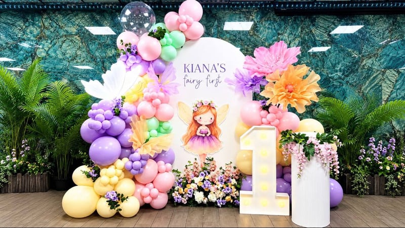 Pastel balloon garland with giant flowers and a lit number one in front of a tropical leaf wall